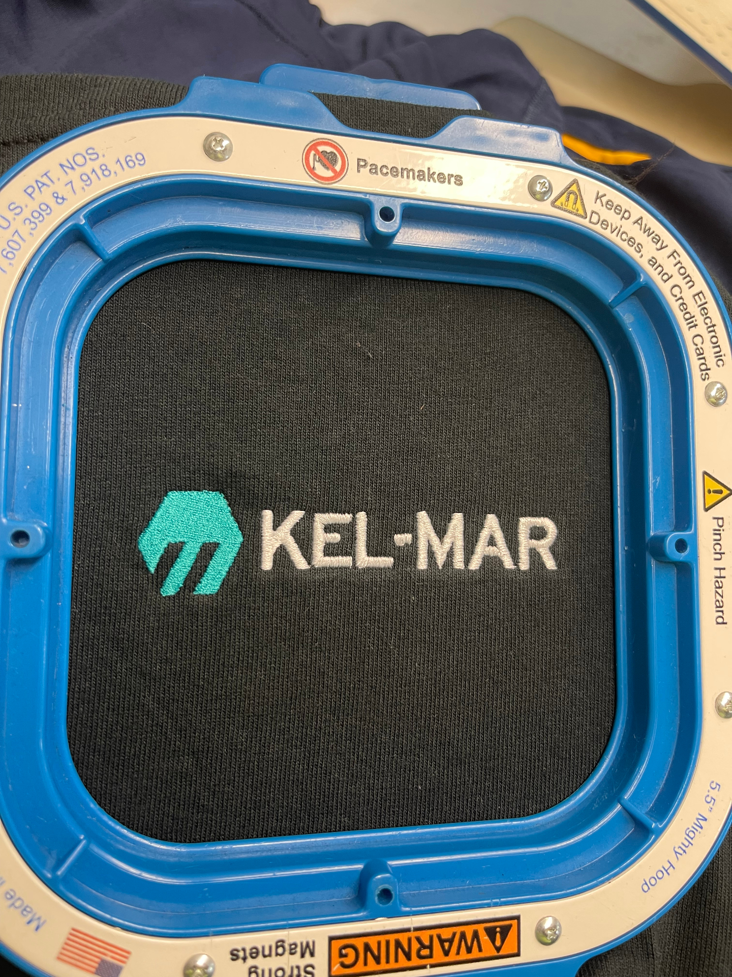 Close-up of Kel-Mar logo embroidery on black apparel in an embroidery hoop.